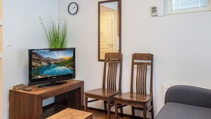 Un salon avec un téléviseur posé sur un meuble en bois, deux chaises en bois, un canapé gris et une petite table en bois.