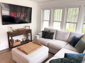 A living room with a sectional sofa, a coffee table, a TV mounted on the wall, a wooden console table, and large windows.
