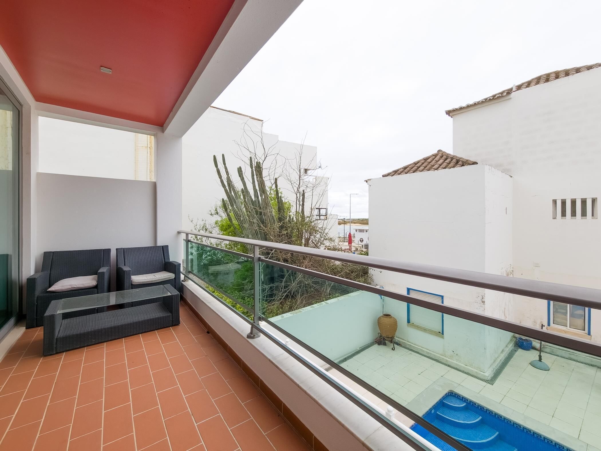 A balcony with a tiled floor, a glass railing, and a view of a swimming pool and buildings.