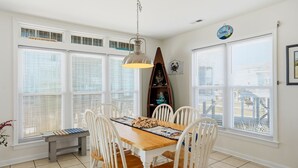 Uma área de refeições com mesa de madeira, cadeiras brancas e uma luminária pendente.