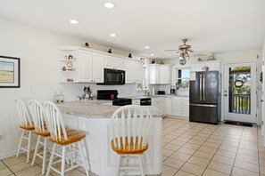 A kitchen with white cabinets, a stainless steel refrigerator, a microwave, and a ceiling fan. There are three white chairs with wooden seats. - 4-bedroom house across from Ocean and Ocean Crest pierwith water view (Oak Island)