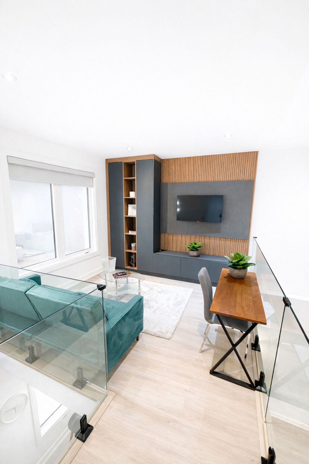 A modern living room with a glass coffee table, teal sofa, wooden accent wall, and a TV mounted on the wall.