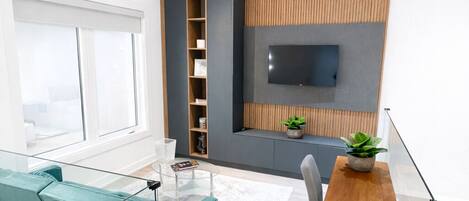 A modern living room with a glass coffee table, teal sofa, wooden accent wall, and a TV mounted on the wall.