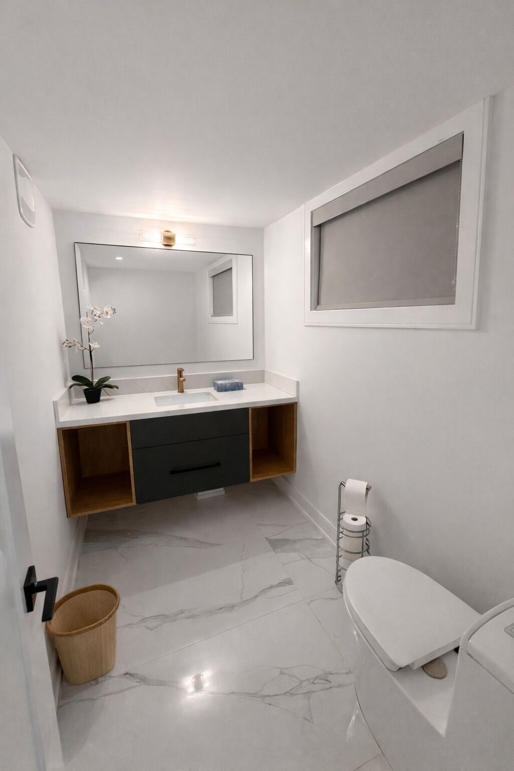 Un baño moderno con un lavamanos, espejo y retrete blancos. Hay una planta pequeña en la encimera del lavamanos y un cubo de basura junto al retrete.