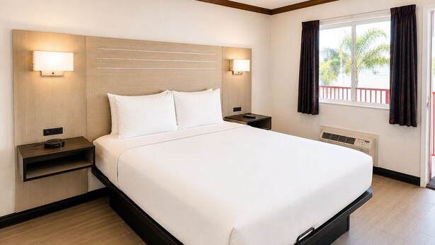 Una habitación de hotel con cama, mesitas de noche, lámparas de pared, una ventana con cortinas y vistas a palmeras.