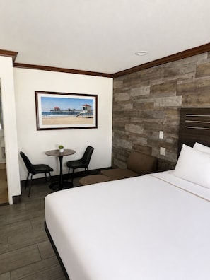 A hotel room with a bed, a small table with chairs, a framed picture on the wall, and a stone accent wall. - Kiwi Inn (Huntington Beach)