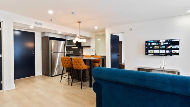 A modern living room with a blue sofa, a kitchen area with a stainless steel refrigerator, and a flat-screen TV mounted on the wall. - Elegant Historic Suite | Two Floors of Luxury (Montreal)