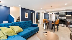 A modern living room with a blue sofa, a round coffee table, and a kitchen area featuring a stainless steel stove and microwave. - Elegant Historic Suite | Two Floors of Luxury (Montreal)