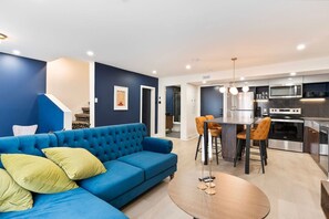 A modern living room with a blue sofa, a round coffee table, and a kitchen area featuring a stainless steel stove and microwave. - Elegant Historic Suite | Two Floors of Luxury (Montreal)
