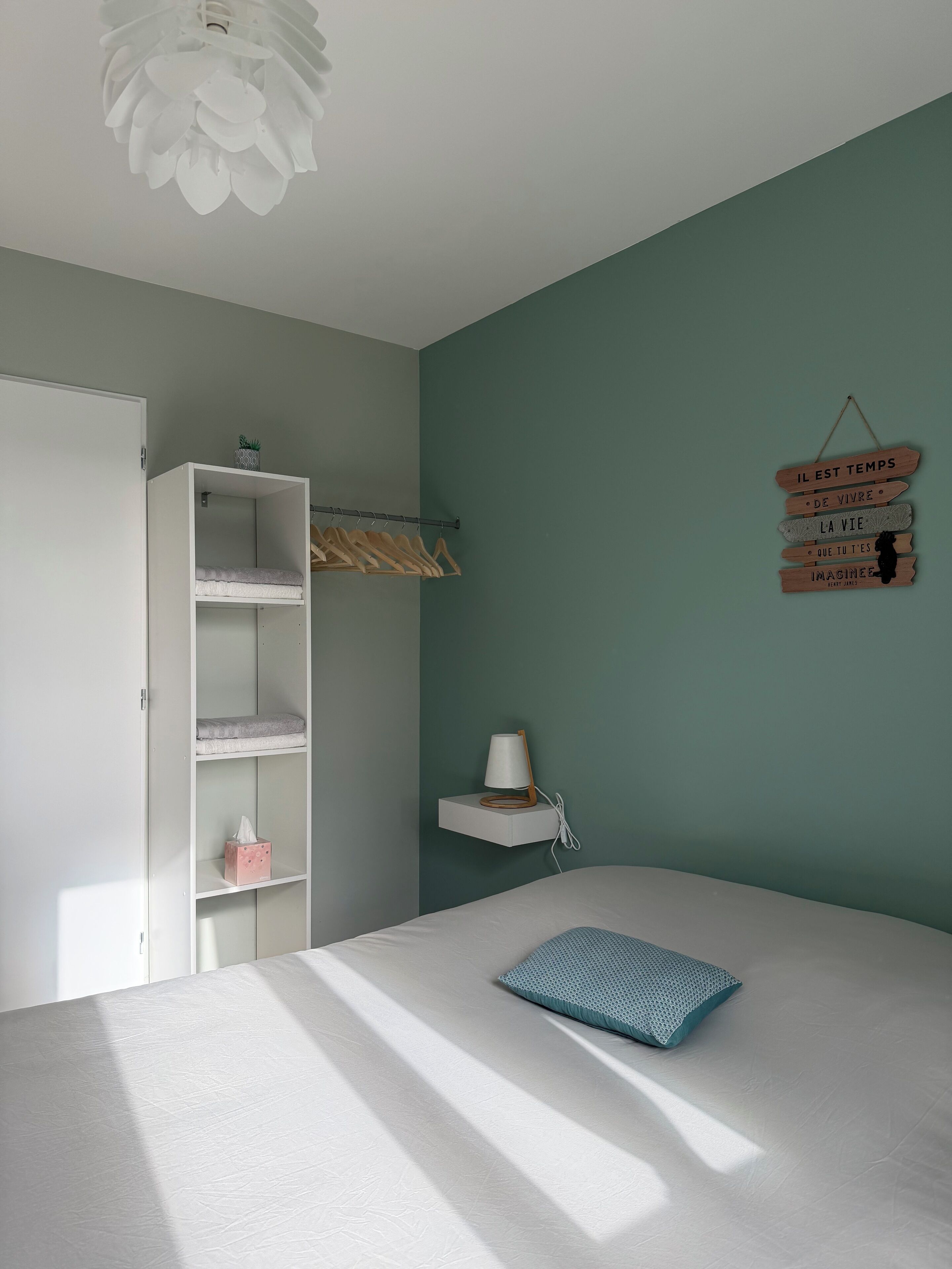 A bedroom with a bed, a white wardrobe, a bedside table with a lamp, and a wall-mounted wooden sign.