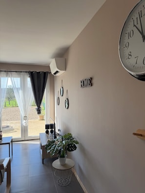 Een kamer met een grote wandklok, een 'HOME' bord, een potplant en uitzicht door een glazen deur.