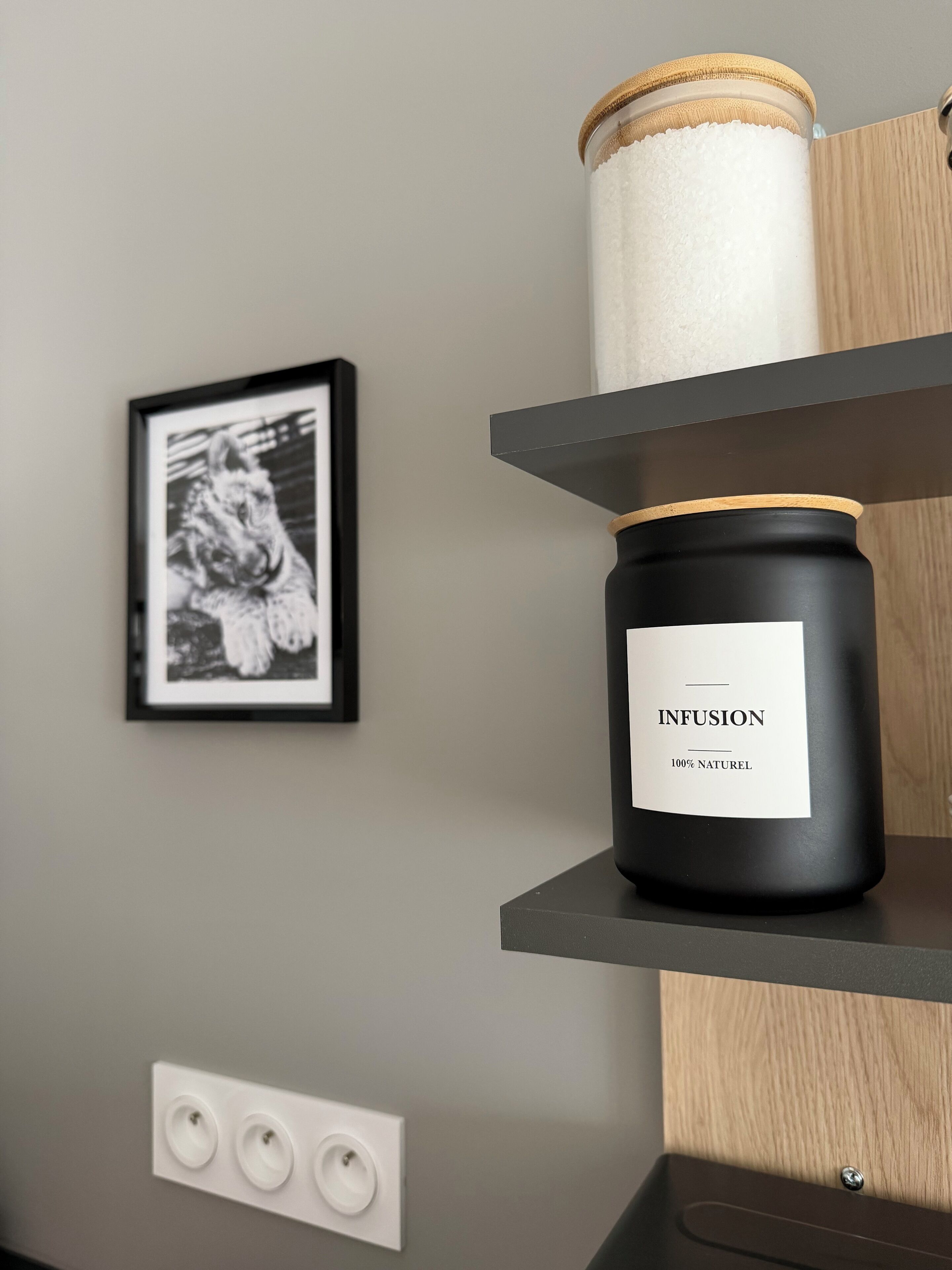 Une bougie noire portant l’inscription « INFUSION » est posée sur une étagère, un tableau encadré est accroché au mur, et un interrupteur se trouve en dessous.