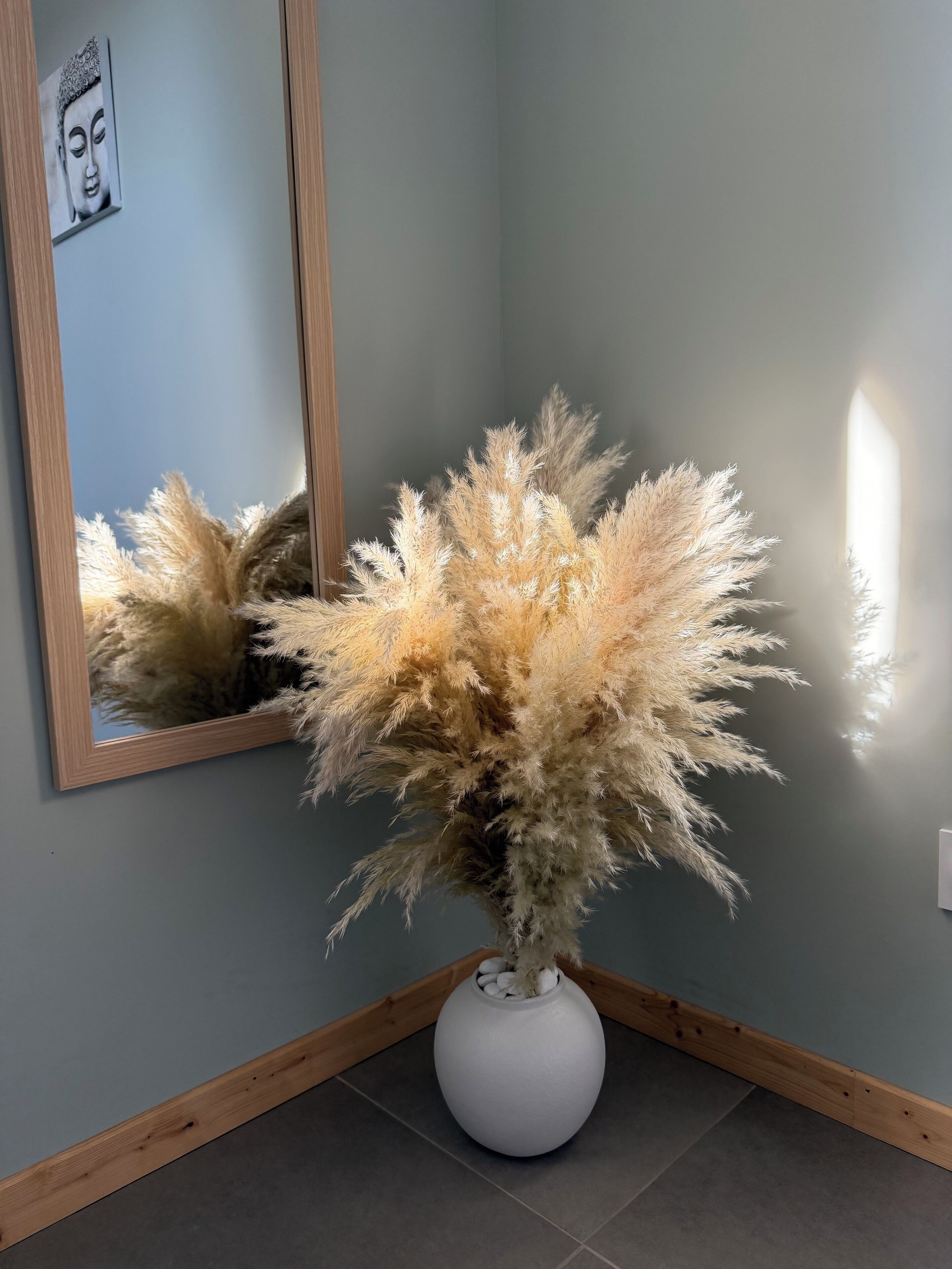 Een kamer met een grote spiegel die een aan de muur bevestigd Boeddhabeeld, een witte vaas met pluimenhalm en een houten vloer weerspiegelt.
