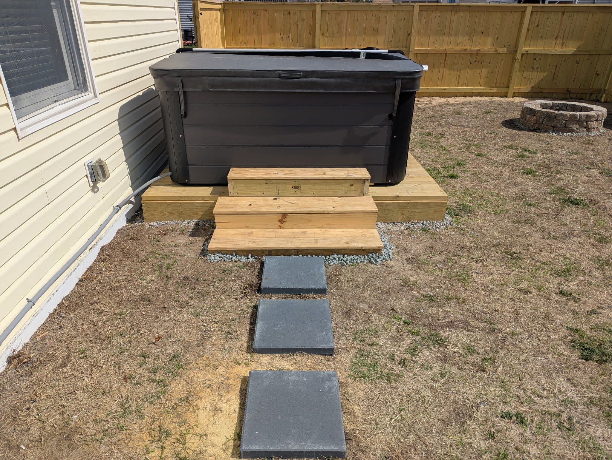 A hot tub with wooden steps leading up to it, placed in a backyard with a wooden fence and a fire pit in the background.