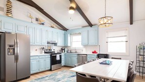A modern kitchen with light blue cabinets, stainless steel appliances, and a large dining table. - New Listing! Walk to Beach, Hot Tub & Games! (Kill Devil Hills)