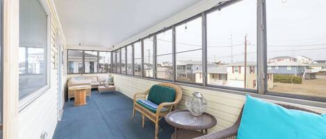 A covered porch with wicker furniture, a small table, and a view of houses in the background.