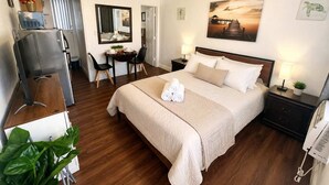 Une chambre d’hôtel avec un lit, un bureau, une chaise, un ventilateur de plafond, un tableau et une plante.