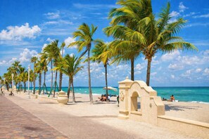 A beach with palm trees, a paved walkway, and a stone archway. - Beachside Studio Steps from the Ocean | Ocean Hollywood Suites (Hollywood)