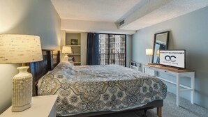 Ein Schlafzimmer mit Bett, Nachttischen, einer Lampe, einem Fenster mit Vorhängen, einem an der Wand befestigten Fernseher und einem gerahmten Schild.
