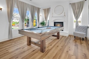 A pool table in a room with wooden flooring, large windows with curtains, a fireplace, and a grey armchair.