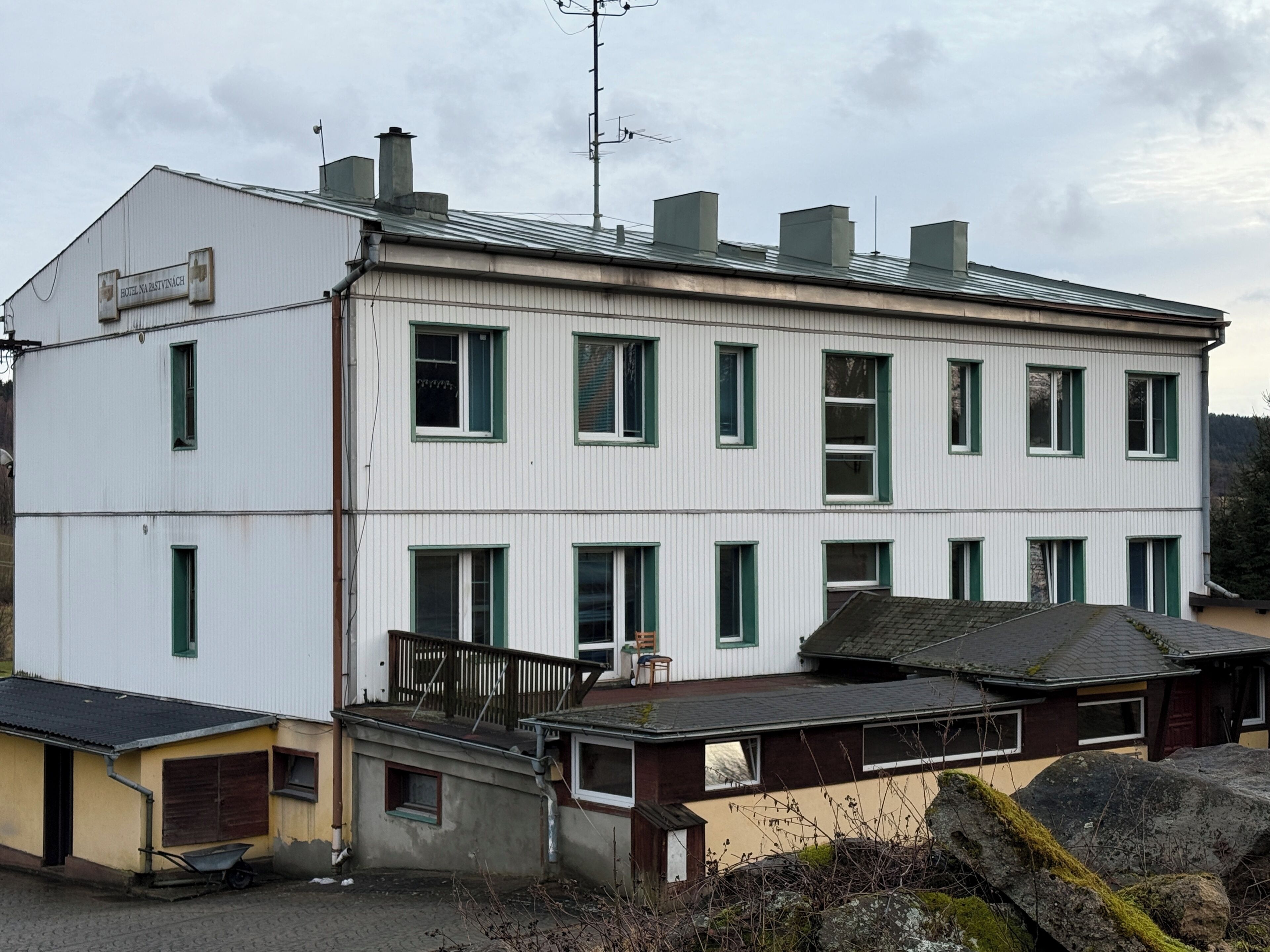 Un edificio de dos pisos con un letrero que indica que es un hotel. Cuenta con varias ventanas con marcos verdes, un balcón y un pequeño espacio de asientos al aire libre.