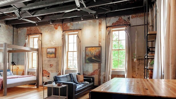 A rustic room with exposed brick walls, a loft bed, a black leather sofa, a wooden dining table, and large windows. - Romantic Historic Loft on Main Street (Heber Springs)
