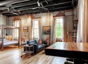 Une pièce au style rustique avec des murs en briques apparentes, un lit en mezzanine, un canapé en cuir noir, une table à manger en bois et de grandes fenêtres.