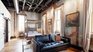 A room with a bed, a black leather sofa, a small table, and a window with curtains. - Romantic Historic Loft on Main Street (Heber Springs)