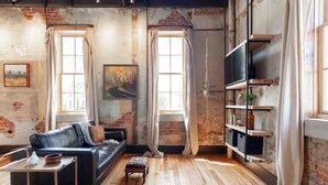 A rustic living room with exposed brick walls, a leather sofa, a coffee table, and a bookshelf. - Romantic Historic Loft on Main Street (Heber Springs)