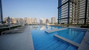 A swimming pool with lounge chairs and umbrellas, surrounded by high-rise buildings. - Dubai Hills Apt with Infinity Pool (Dubai)