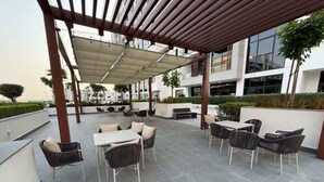 Ein Außenbereich mit Terrassentischen und -stühlen, einer Pergola, die Schatten spendet, und einem modernen Gebäude im Hintergrund.