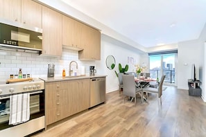 A modern kitchen with wooden cabinets, a built-in oven, and a dishwasher. There is a dining area with a table and chairs, and a view of the city through large windows. - New High Rise Building w Gorgeous Views (Kitchener)