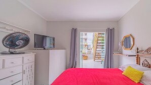 Een slaapkamer met een bed, een televisie, een ventilator, een dressoir en uitzicht op een balkon met planten.