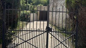 Une entrée fermée par un portail mène à un bâtiment à la façade en pierre. Un chemin pavé conduit à l’entrée, et un paysage vallonné se dessine à l’arrière-plan.