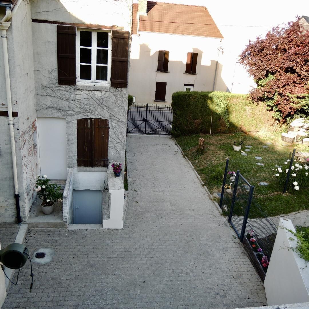 Une cour intérieure avec un chemin pavé menant à une entrée fermée par un portail, le tout entouré de bâtiments aux murs blancs et aux volets sombres.