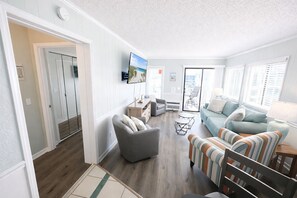 A spacious living area with a sofa, armchair, and a television mounted on the wall. - Ocean View Condo Steps from the Beach (Myrtle Beach)