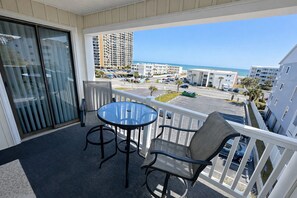 A balcony with a table and chairs, overlooking a parking area and buildings with a view of the beach. - Ocean View Condo Steps from the Beach (Myrtle Beach)