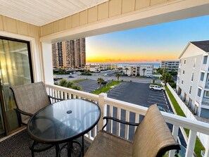 A balcony with a glass table and chairs, overlooking a parking lot and buildings with a sunset view. - Ocean View 2BR + Steps from the Beach + Pool (Myrtle Beach)
