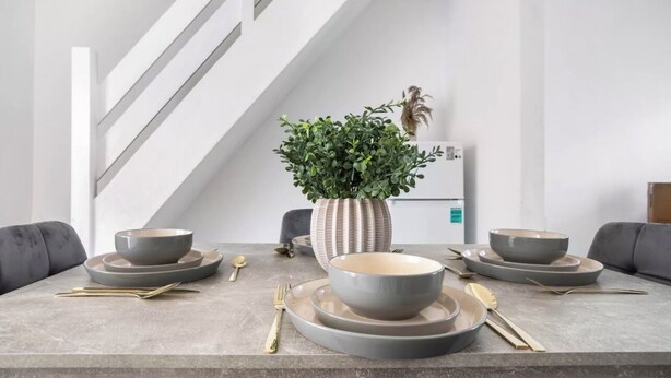 A modern dining area with a grey table set for four, a potted plant, and a refrigerator in the background.