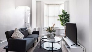 A modern living room with a dark grey sofa, a glass coffee table, a flat-screen TV on a white stand, and a large window with sheer curtains.