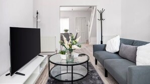 A modern living room with a flat-screen TV, a round glass coffee table with a vase of flowers, a grey sofa, and a coat rack.