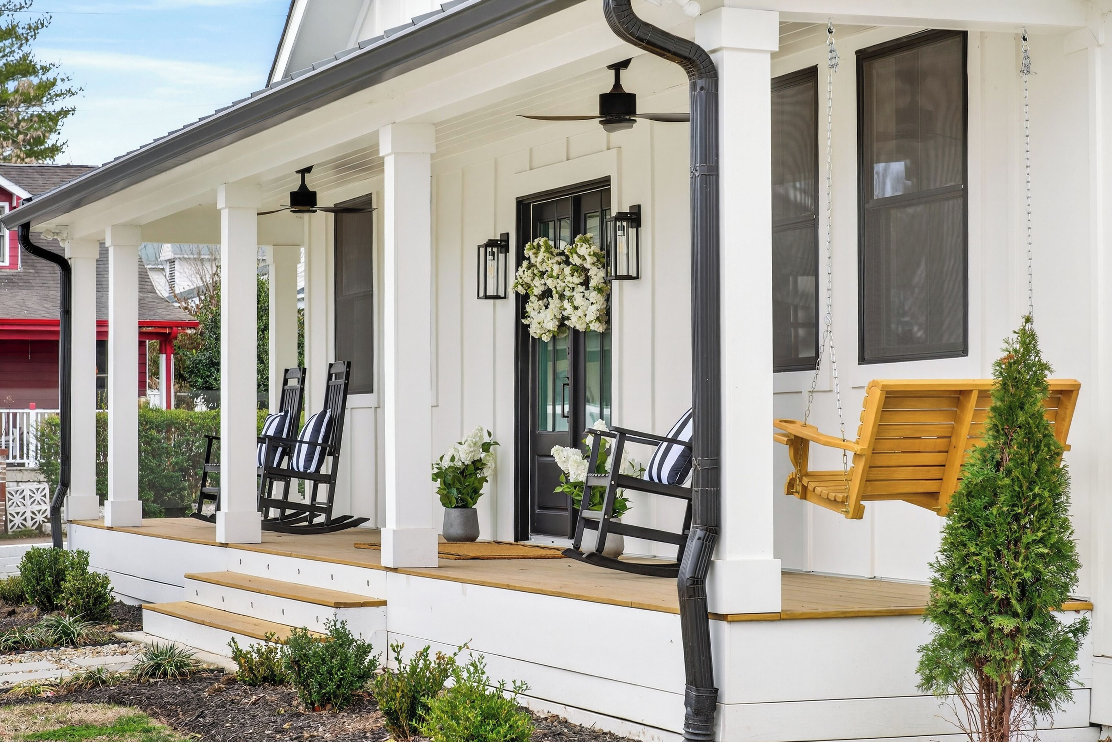 A white porch with black-framed chairs and a wooden swing, surrounded by small plants and a potted tree.