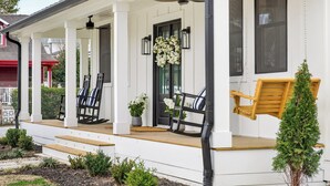 A white porch with black-framed chairs and a wooden swing, surrounded by small plants and a potted tree. - Luxury Franklin Retreat • Walk to Downtown • Front Porch (Franklin)