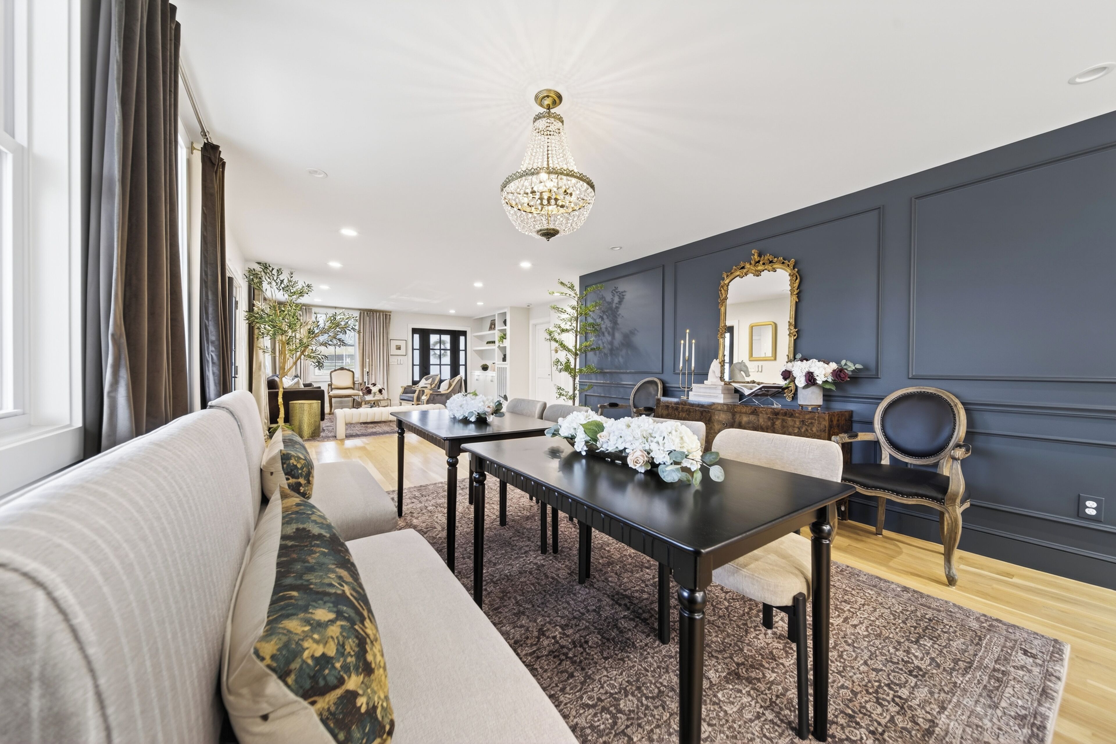 Un salon moderne avec un mur d’accent de couleur foncée, un lustre, une table à manger agrémentée de compositions florales et un grand miroir.
