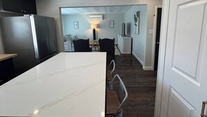 A modern kitchen with a white marble countertop and a stainless steel refrigerator. There is a dining area with a table and chairs, and a hallway leading to a living space.