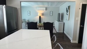 A modern kitchen with a marble countertop, a dining area with a table and chairs, a TV, and a hallway leading to another room.