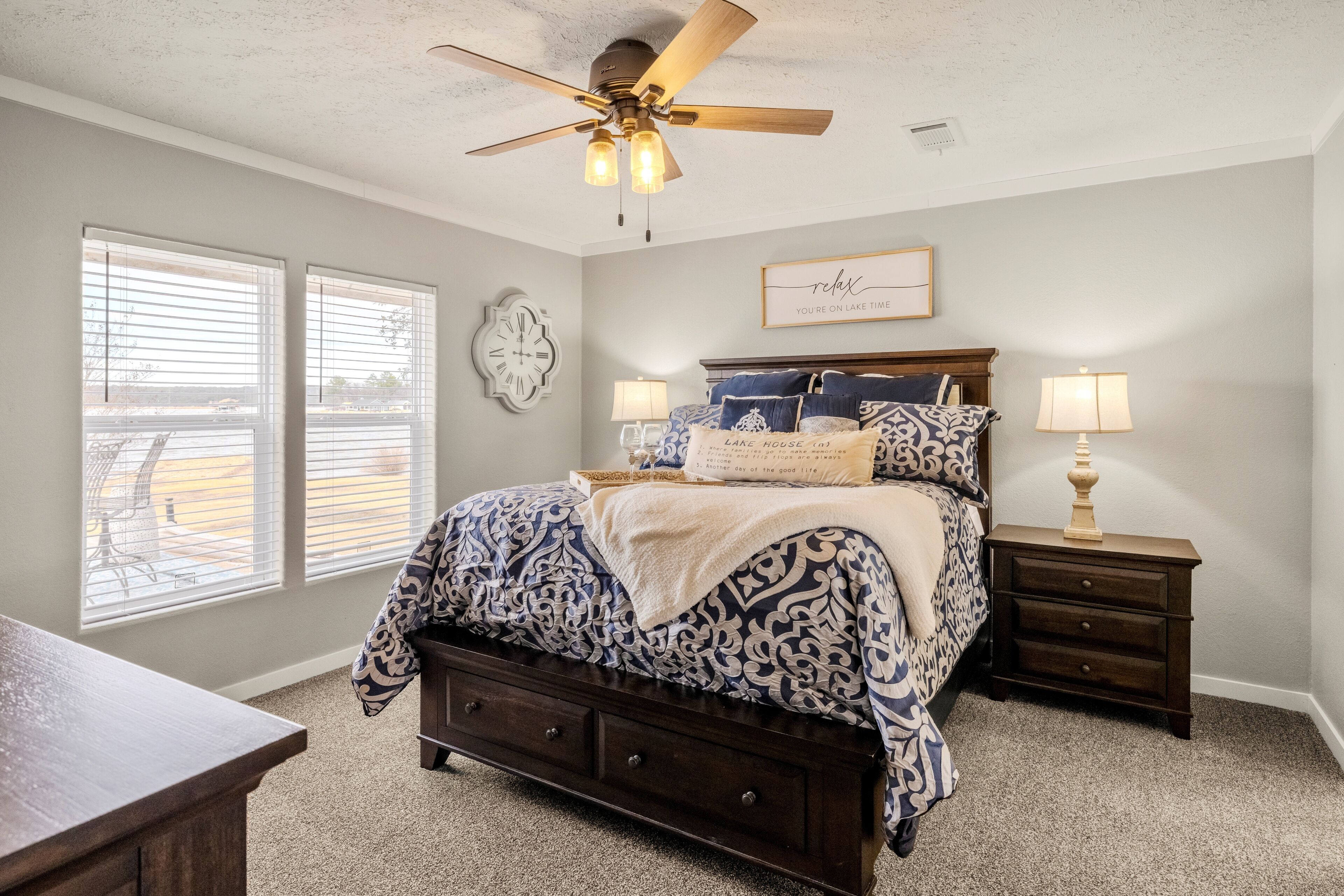A bedroom with a bed, two bedside tables, a ceiling fan, a clock, and a window with blinds.