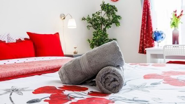 A neatly made bed with a floral-patterned bedspread, a red pillow, and a grey towel rolled up. - Modern 1-bed in West Kensington Near Tube (London)