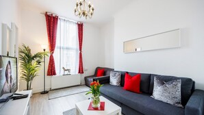 A modern living room with a grey sofa, red and white cushions, a white coffee table with a vase of flowers, a TV on a white stand, and a large window with red curtains. - Modern 1-bed in West Kensington Near Tube (London)