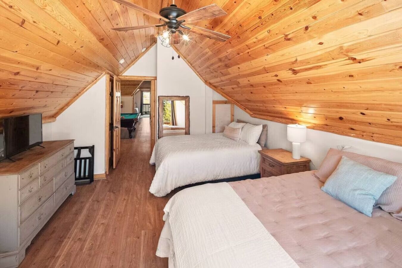 A loft-style bedroom with wooden beams, two beds, a ceiling fan, a dresser, and a television.